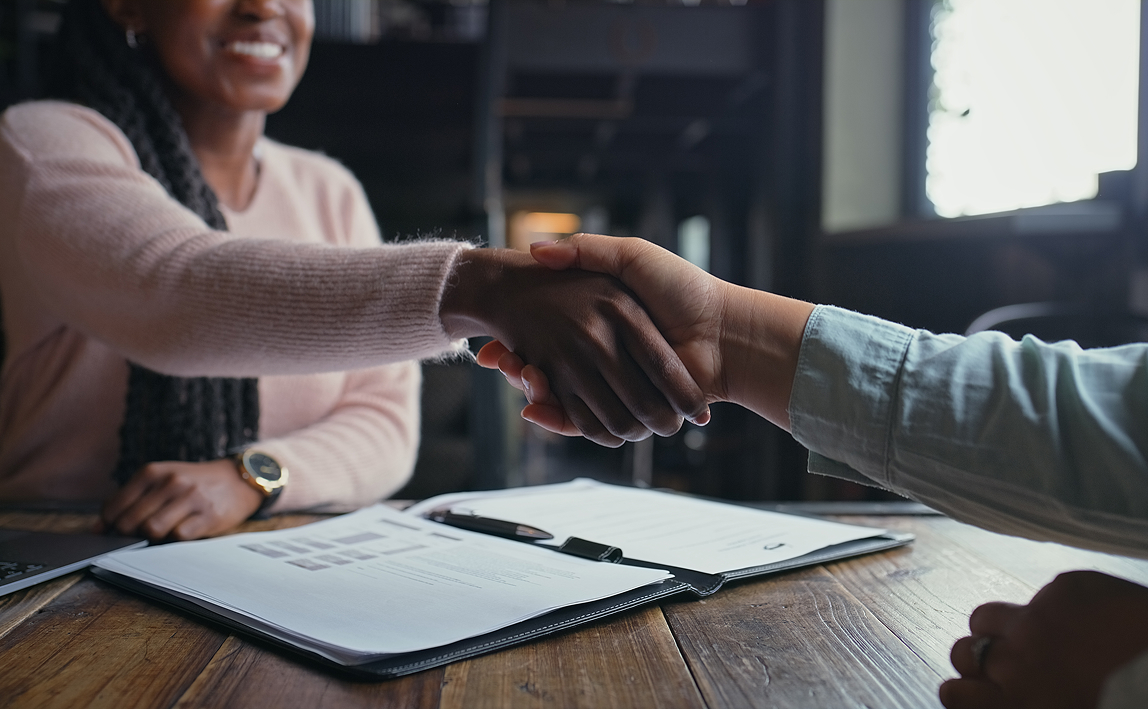 Business people, handshake and documents with meeting for interview, hiring or recruiting at cafe. Closeup, employer and candidate shaking hands for career opportunity or partnership at coffee shop